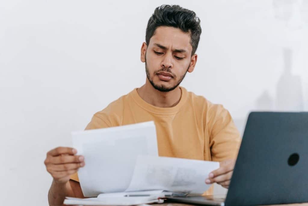 How to Deal with Medical Bills in Collections: Steps to Take & Options 3 Young man in a yellow shirt reviewing paperwork with a concerned expression beside a laptop