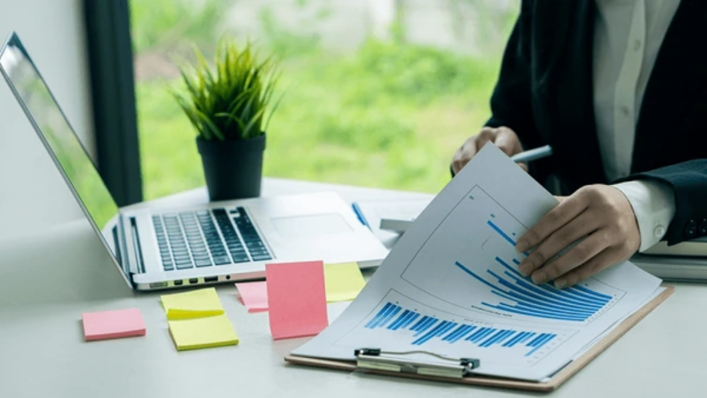 Business professional at desk reviewing financial charts and data on clipboard with laptop and colorful sticky notes