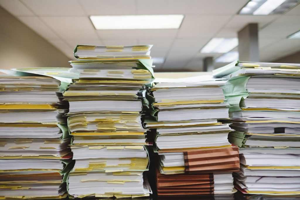 Multiple tall stacks of papers and documents with colored tabs and folders piled on an office desk