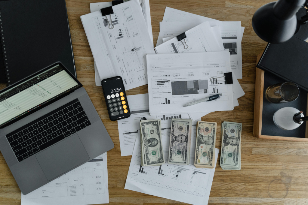 Overhead view of a wooden desk with a laptop, a smartphone, financial reports, US dollar bills, and a coffee cup arranged on the workspace