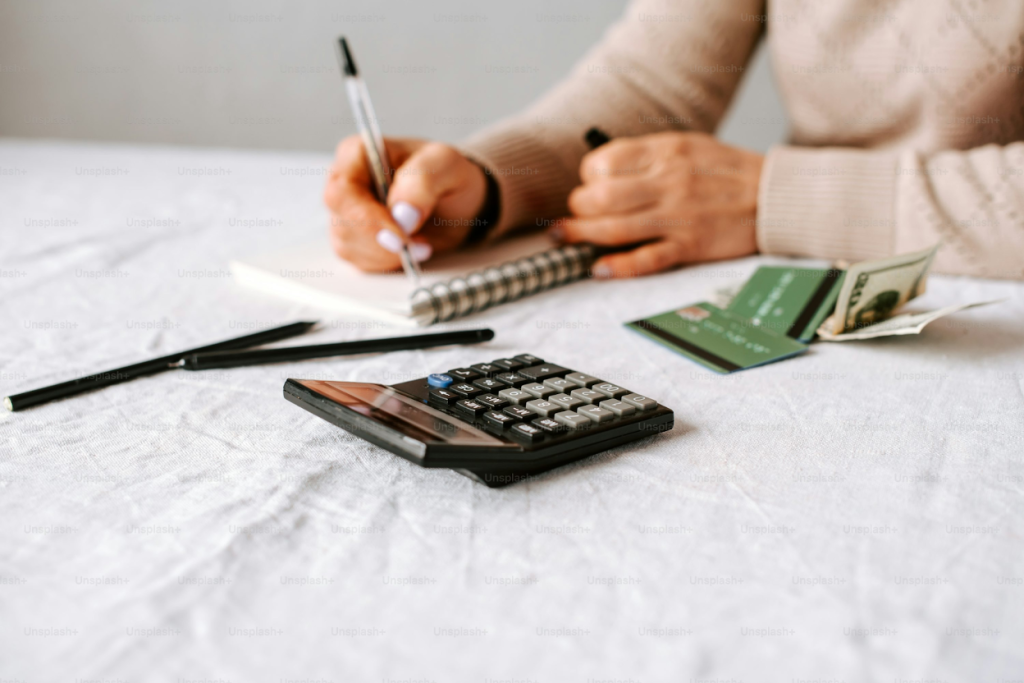 Person writing in a notebook at a desk with a calculator, credit cards, cash, and financial documents spread out on the table