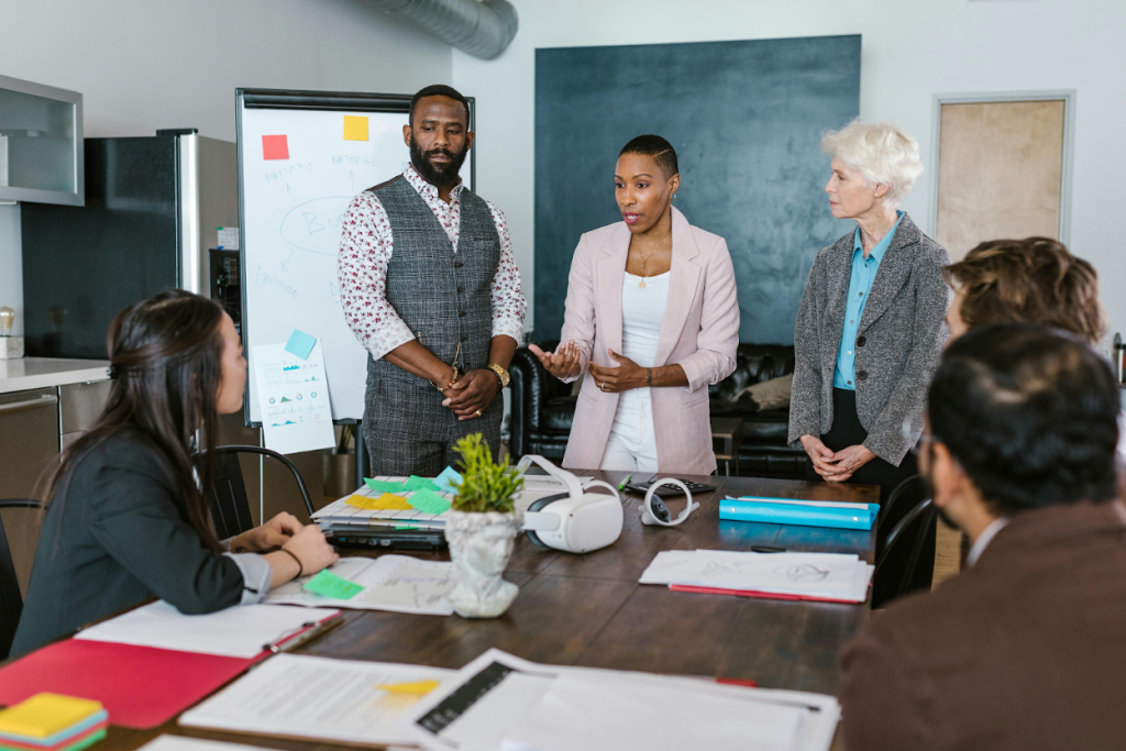 Professionals in a conference room meeting, with three presenters standing and addressing colleagues seated around a conference table.