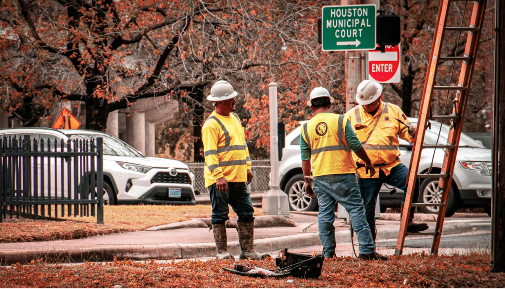 Three municipal workers wearing yellow safety vests and white hard hats are working on infrastructure near a street