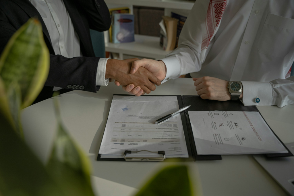 Two business professionals in formal attire shaking hands across a desk