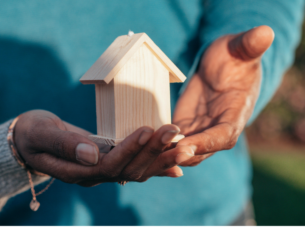 Two hands holding a small wooden house model against a teal background