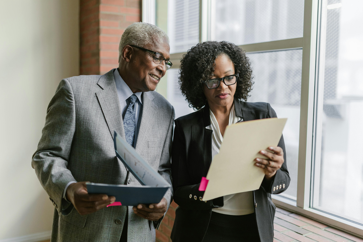 Two professionals in business suits review paperwork together by an office window