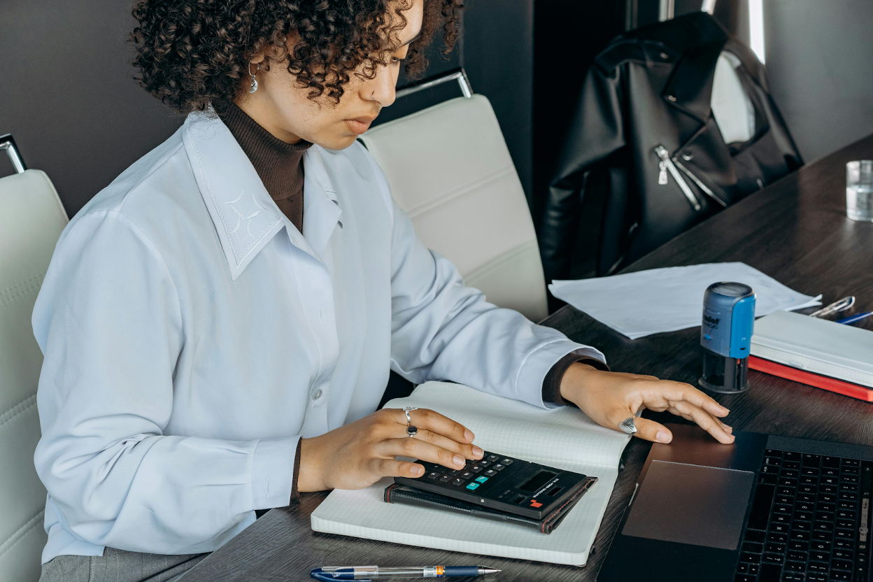Woman in a white blazer using a calculator while reviewing documents at a desk with a laptop