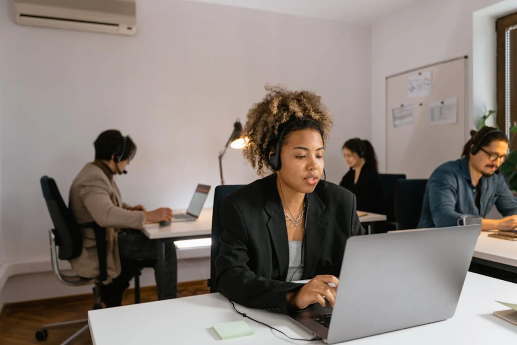 Professional woman in a black blazer working on a laptop in a modern office with colleagues in the background