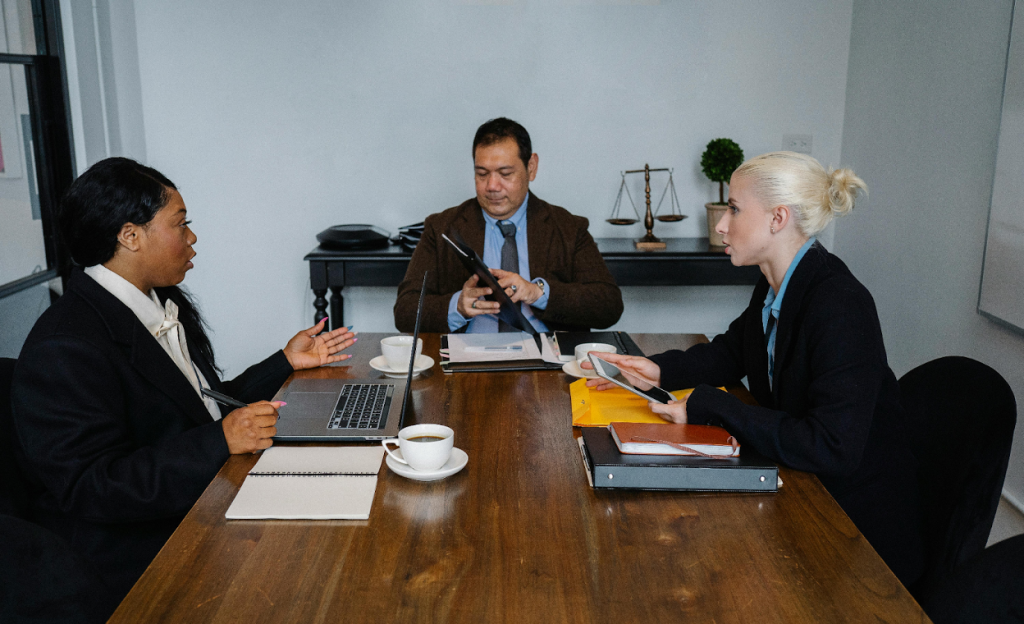 Three business professionals are having a meeting around a wooden conference table.