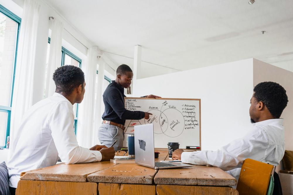 Three professionals in white shirts gathered around a table with a laptop, one standing and presenting diagrams on a whiteboard during a business meeting.