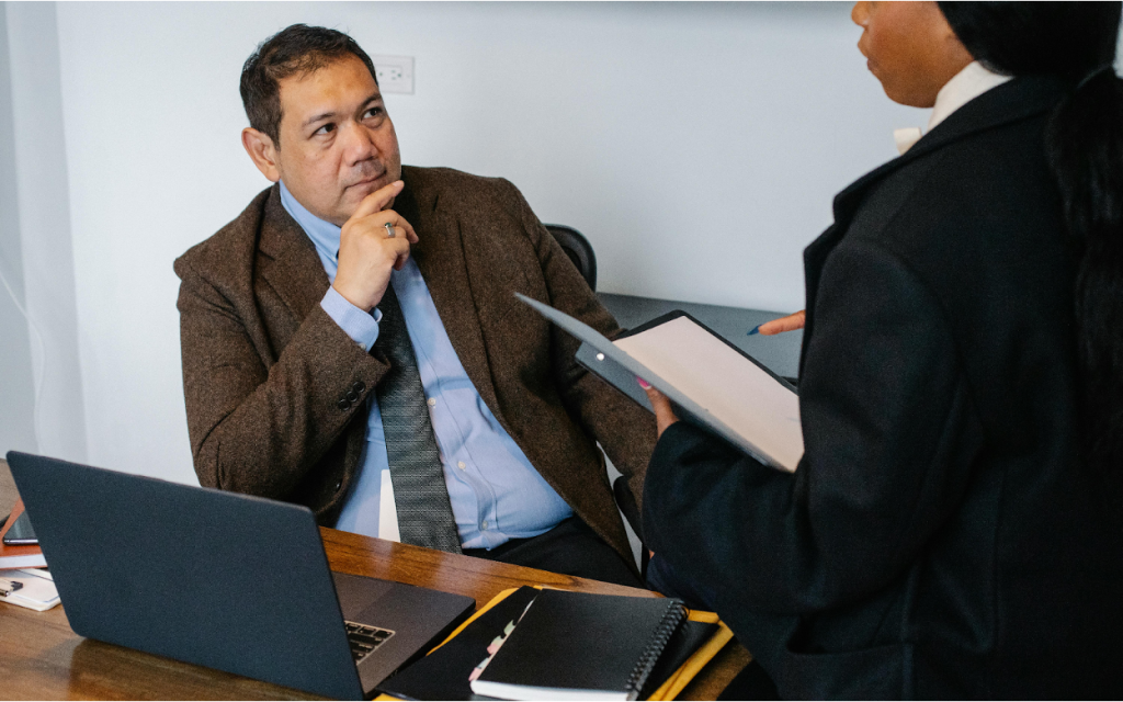 Business professional in brown blazer sitting at desk, listening thoughtfully to colleague