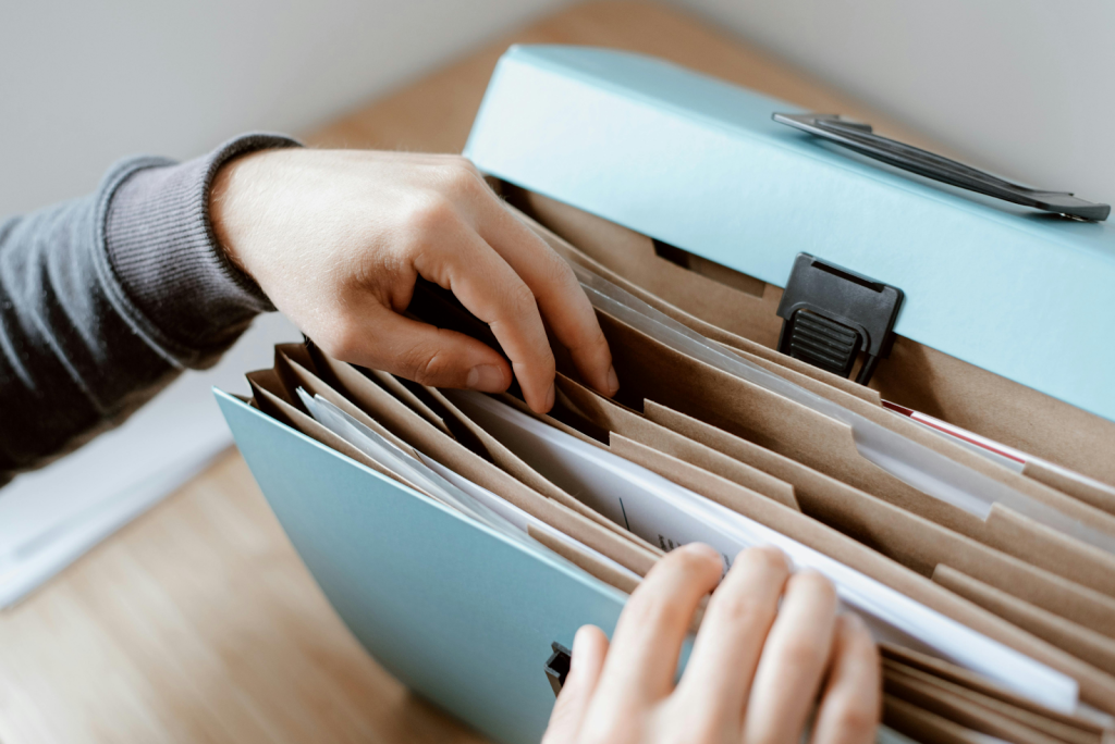 Hands sorting through documents in an expandable accordion file folder on a wooden desk.
