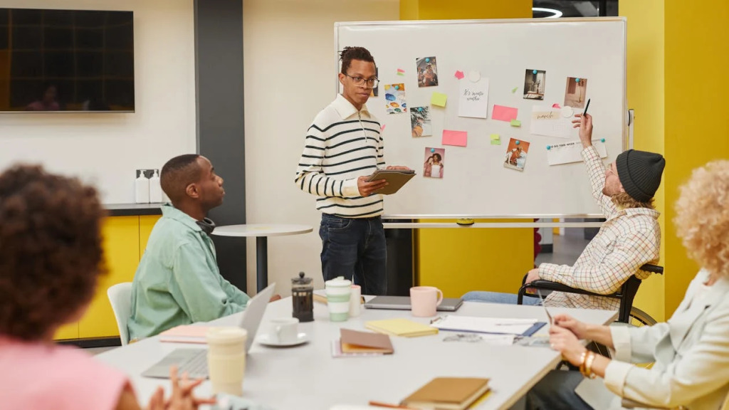 Person in striped sweater presenting at a whiteboard with sticky notes while colleagues listen around a table.