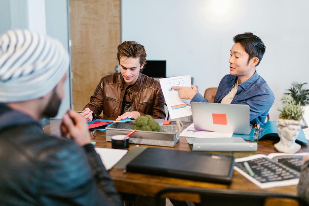 Three business professionals collaborating at a conference table with laptops, documents, colorful charts, and office supplies during a working meeting