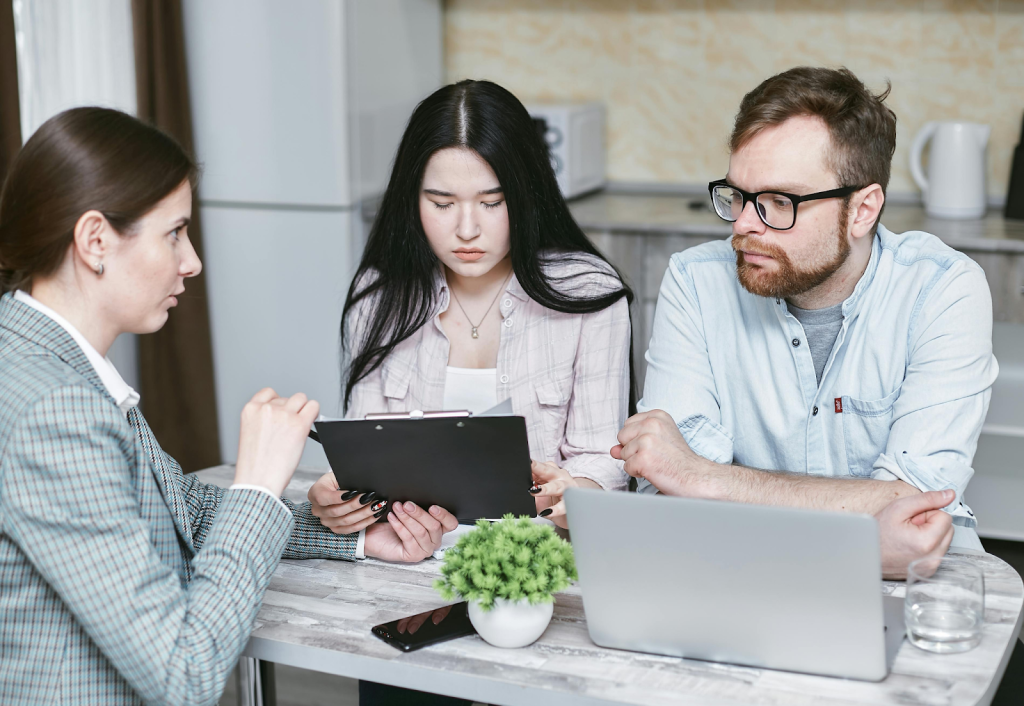 Three professionals in a meeting, reviewing documents on a clipboard together at a table with a laptop.