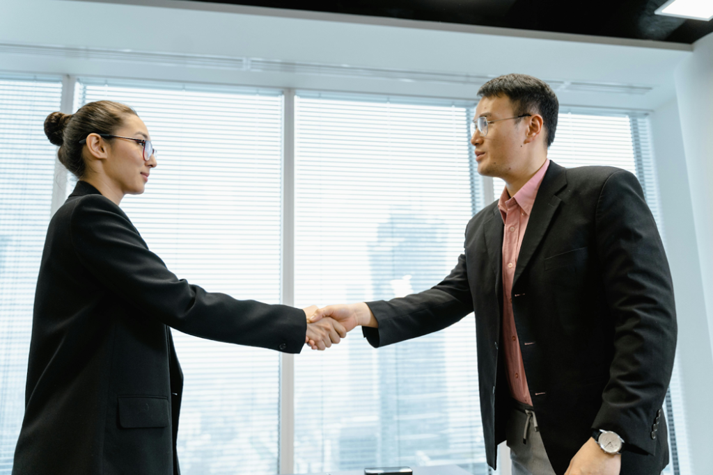 Two business professionals in dark suits shaking hands in a modern office with large windows