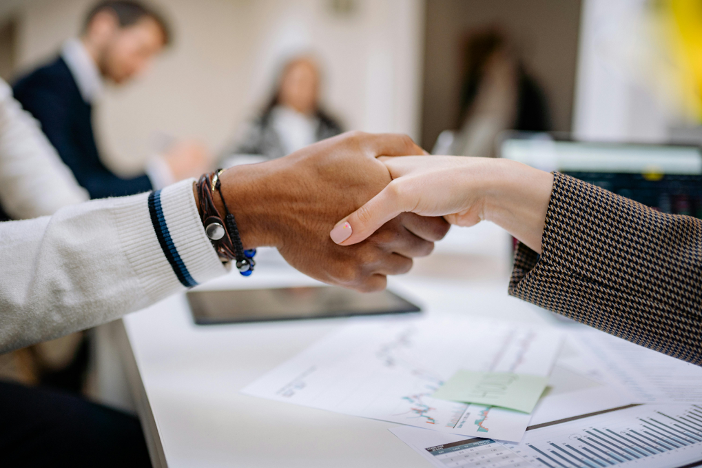 Two business professionals shaking hands across a desk covered with documents and charts in an office setting