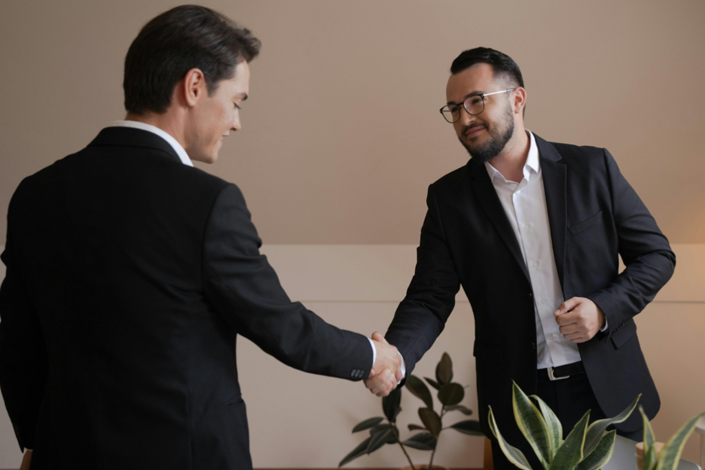 Two businessmen in dark suits shaking hands in a modern office setting with a plant in the background