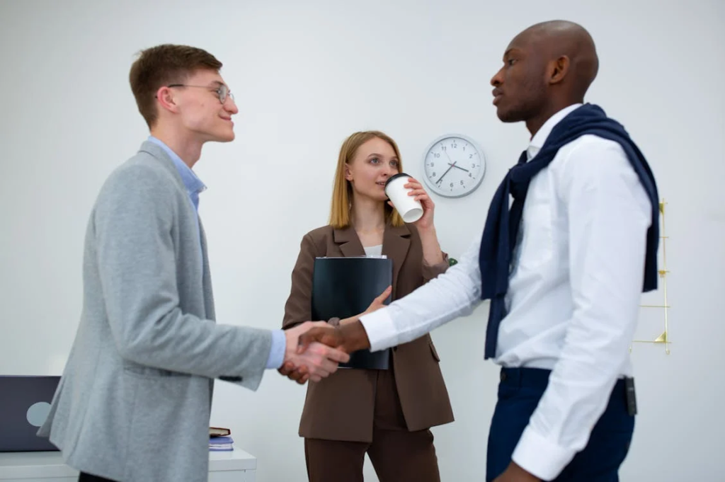 Two businessmen shaking hands in an office while a professional woman holding a folder and a coffee cup stands between them, with a wall clock visible in the background