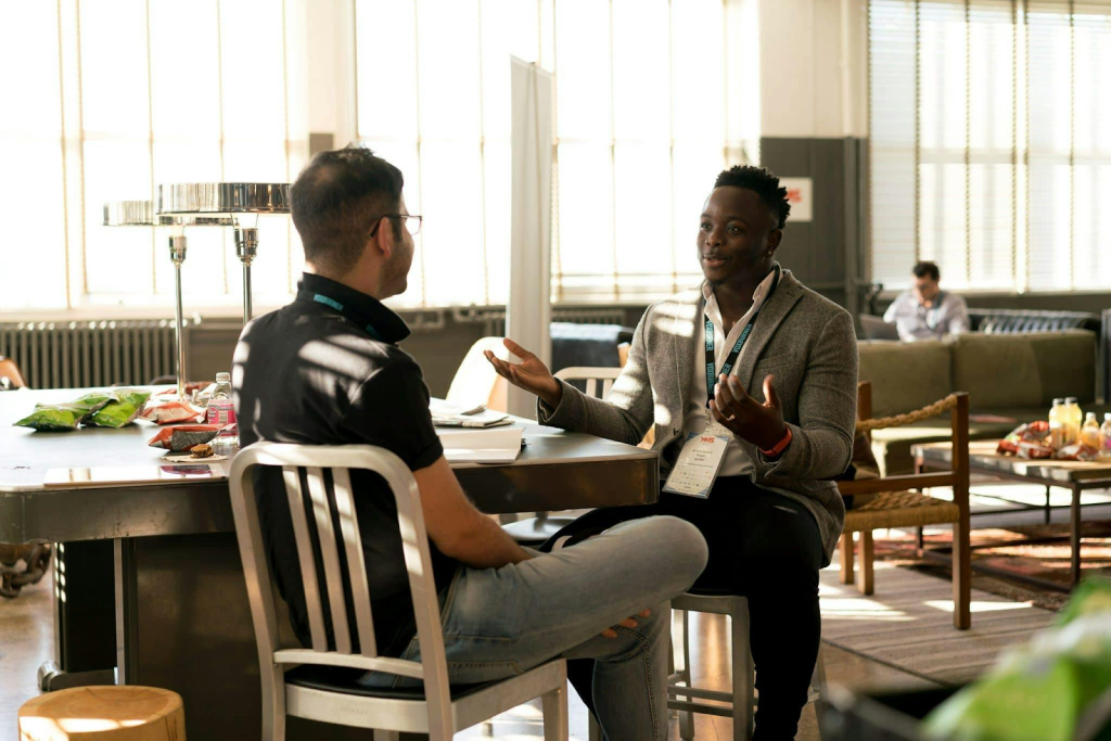 Two men having a conversation at a table during a conference or networking event in a modern space.