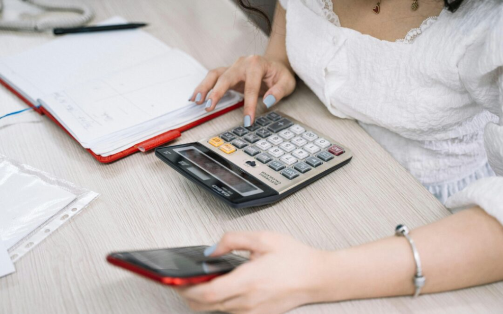 Woman using a calculator while holding a smartphone at a desk with documents.