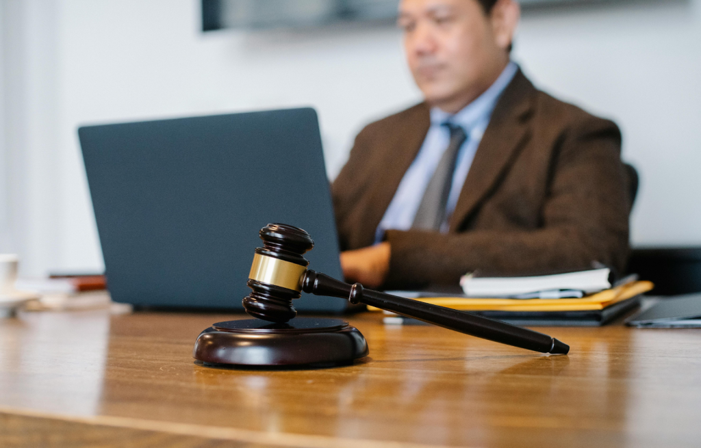 Wooden gavel with gold band on a desk, with a man in a brown suit working on a laptop in the background.