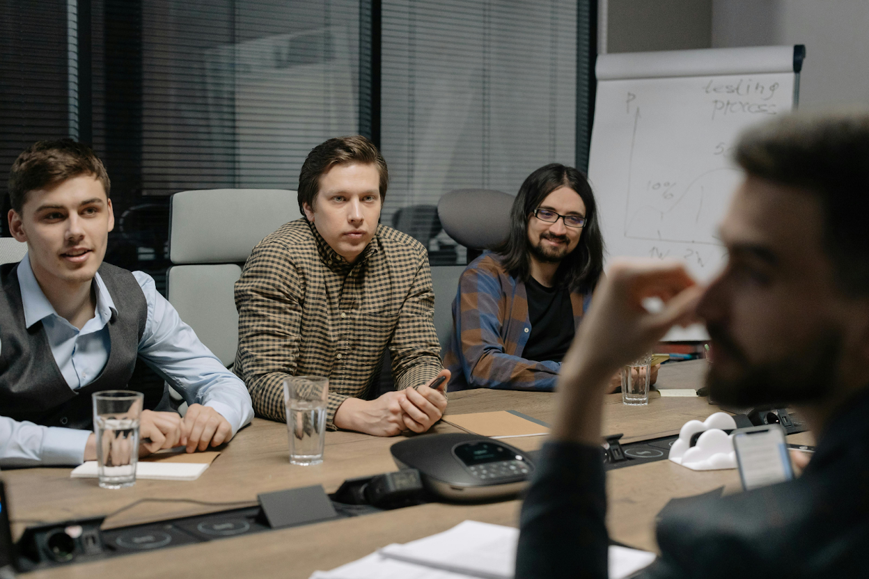 Four men in a conference room meeting with a whiteboard, conference phone, and water glasses on the table