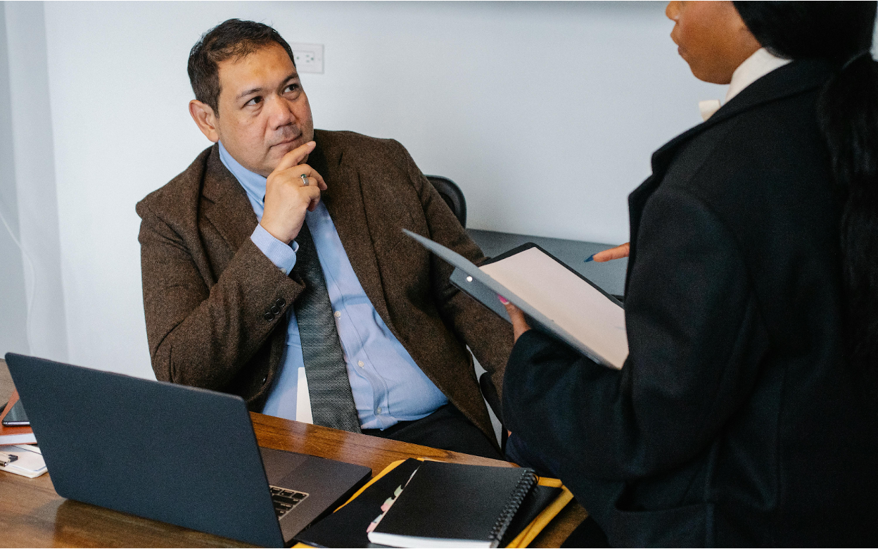 Man in brown suit jacket listening thoughtfully during business meeting while colleague presents documents on tablet