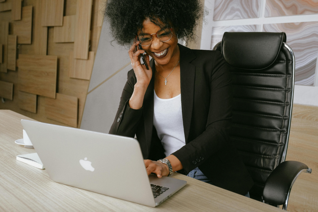 Professional woman in a black blazer smiling while on a phone call at her desk with a laptop