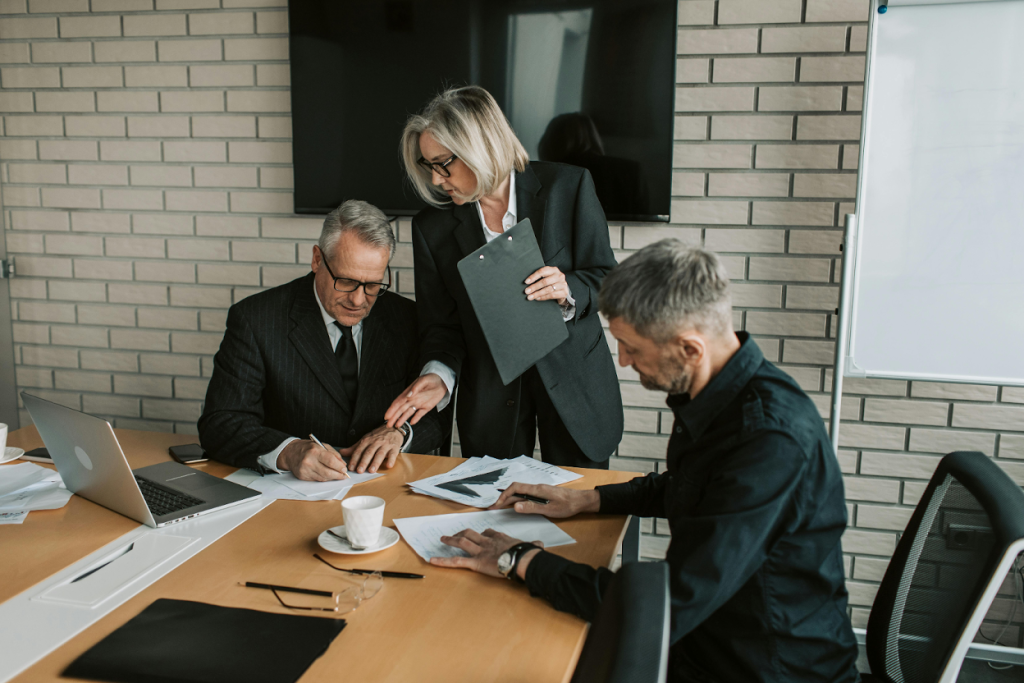 Three business professionals in suits reviewing documents at a conference table with a brick wall background