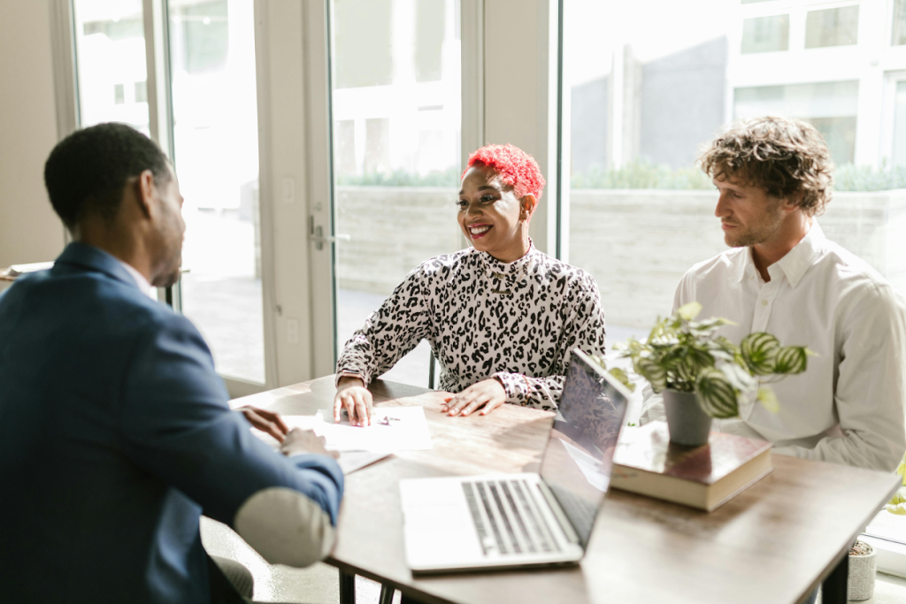 Three professionals in a bright office meeting around a table with laptop and plants