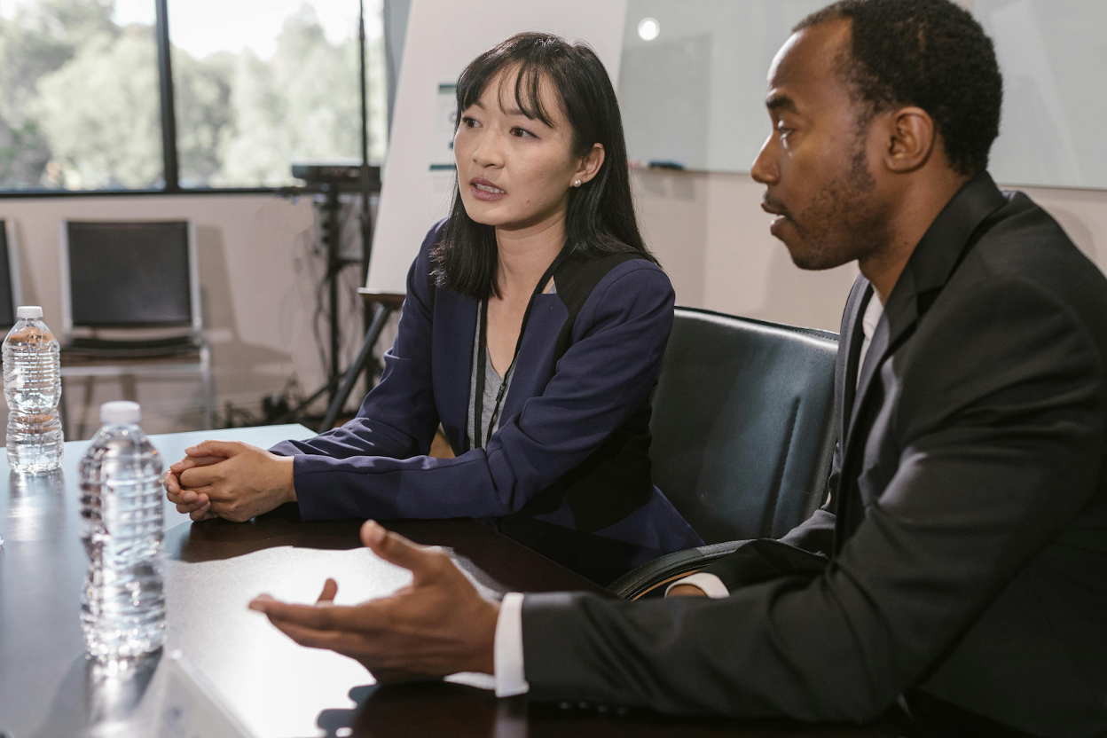 Two business professionals in formal attire engage in a discussion at a conference table in a modern office