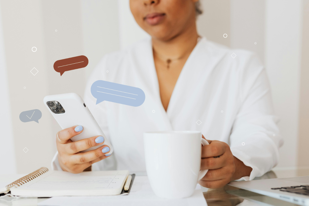 Woman typing on smartphone while holding a mug with documents on a table