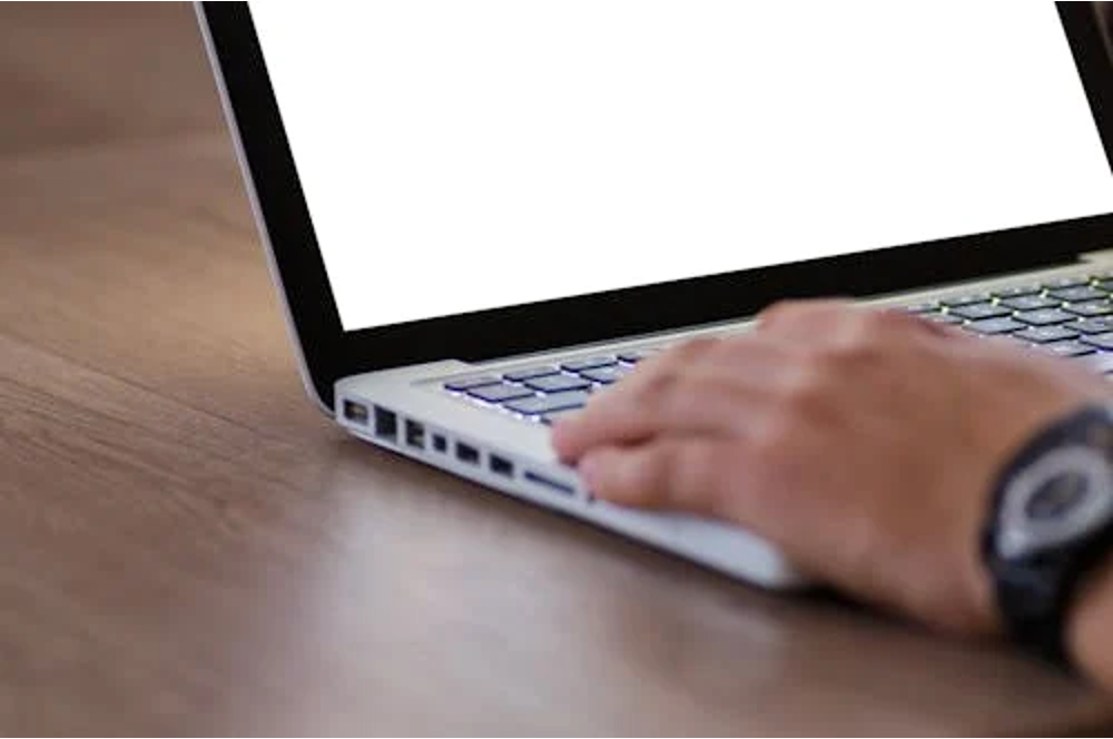 A person's hand typing on a laptop with a blank white screen on a wooden desk