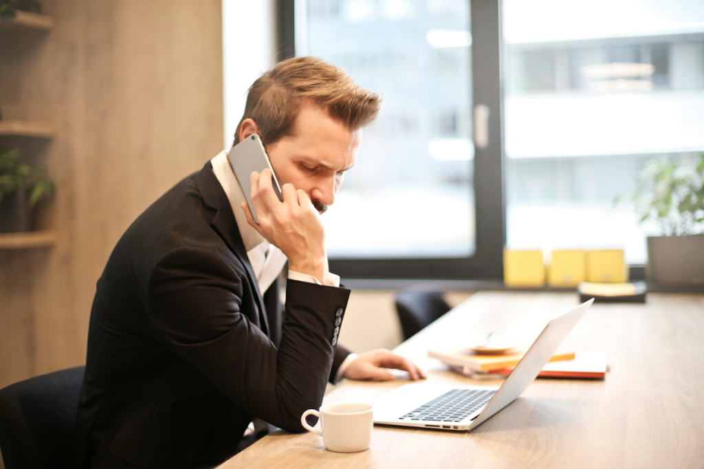 Man in a dark suit talking on a phone while sitting at a desk with a laptop and a coffee cup