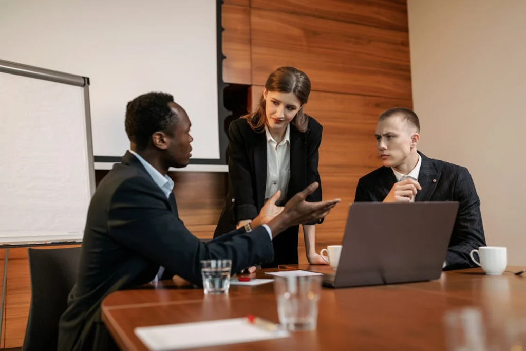 Three business professionals in suits discussing at a conference table with a laptop.