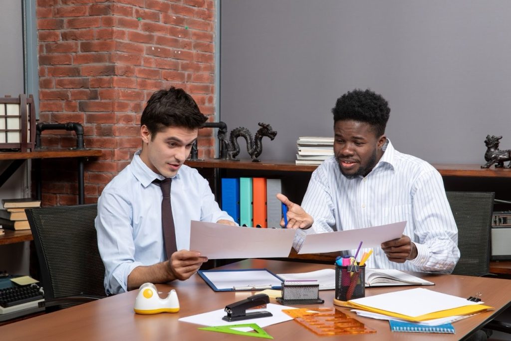 Two business professionals in an office are discussing invoice documents and payment records spread across a conference table
