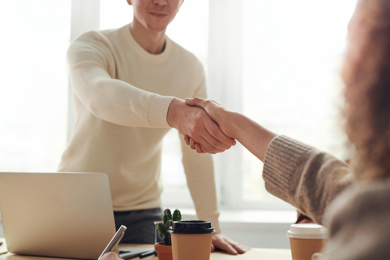 Two people shaking hands across a desk with a laptop, coffee cups, and a small cactus